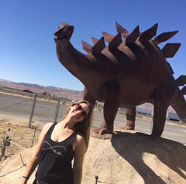 Emma smiles in front of an iron stegosaurus sculpture in the desert.